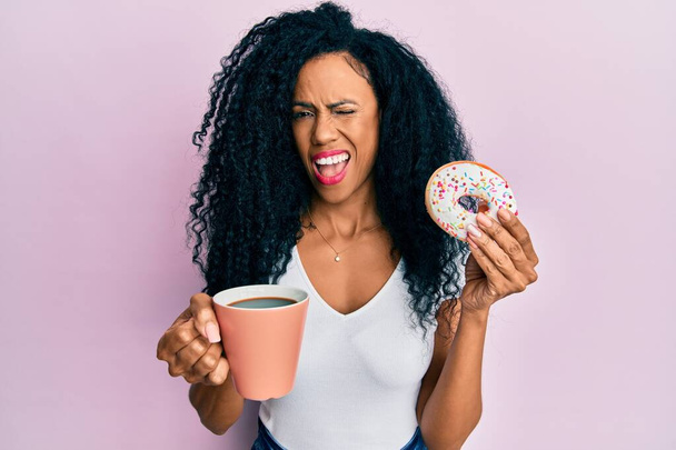 Středního věku africký Američan žena jíst koblihu a pití kávy mrkání na kameru se sexy výraz, veselý a šťastný obličej.  - Fotografie, Obrázek