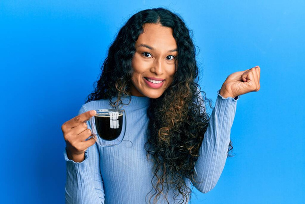 Giovane donna latina che tiene il caffè urlando orgoglioso, celebrando la vittoria e il successo molto eccitato con il braccio alzato  - Foto, immagini