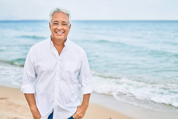 Handosme homme hispanique aux cheveux gris souriant heureux à la plage, profitant de vacances en été - Photo, image