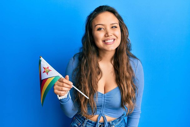 Young hispanic girl holding zimbabwe flag looking positive and happy standing and smiling with a confident smile showing teeth  - Photo, Image