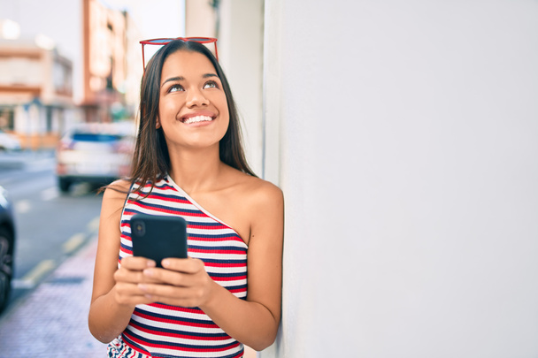 Joven chica latina sonriendo feliz usando smartphone en la ciudad. - Foto, imagen