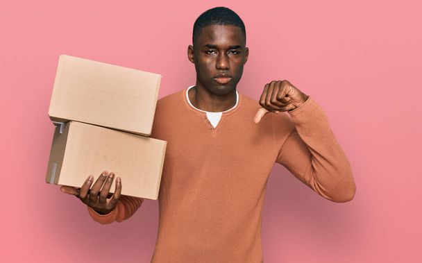 Młody Afroamerykanin trzymający przesyłkę z wściekłą twarzą, negatywny znak pokazujący niechęć z opuszczonymi kciukami, koncepcja odrzucenia  - Zdjęcie, obraz