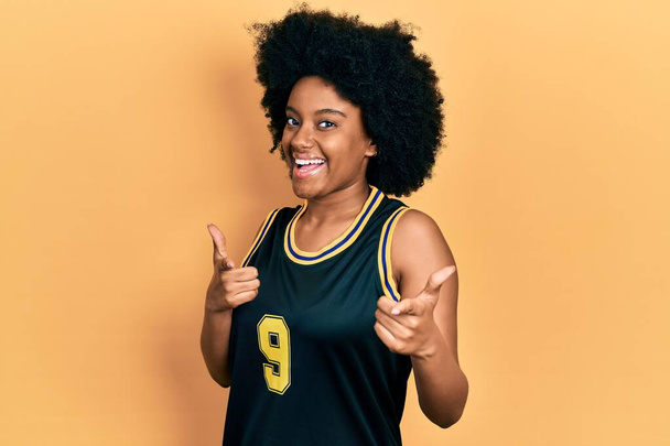 Genç Afro-Amerikan kadın basketbol üniforması giyip kamerayı mutlu ve komik yüzüyle işaret ediyor. iyi enerji ve titreşimler.  - Fotoğraf, Görsel