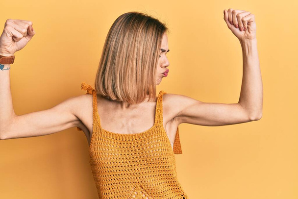 Молодая кавказская блондинка в непринужденной желтой футболке с гордо улыбающимися мышцами рук. фитнес концепция.  - Фото, изображение