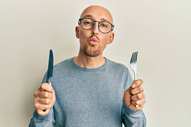 Bald man with beard holding fork and knife ready to eat looking at the camera blowing a kiss being lovely and sexy. love expression.  - Photo, Image