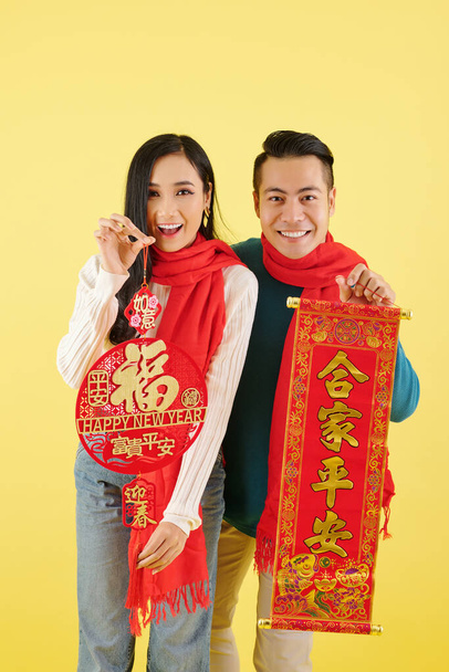 Heureuse jeune femme asiatique excitée dans des vêtements chauds d'hiver montrant des décorations pour le Nouvel An chinois avec l'inscription de meilleurs voeux - Photo, image