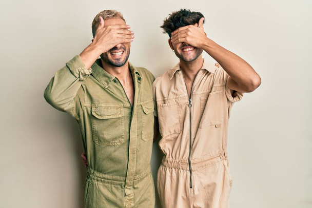 カジュアルなジャンプスーツを着て一緒に立っている同性愛者のゲイのカップルは笑顔と驚きのために目をカバー顔に手で笑っています。視覚障害の概念.  - 写真・画像