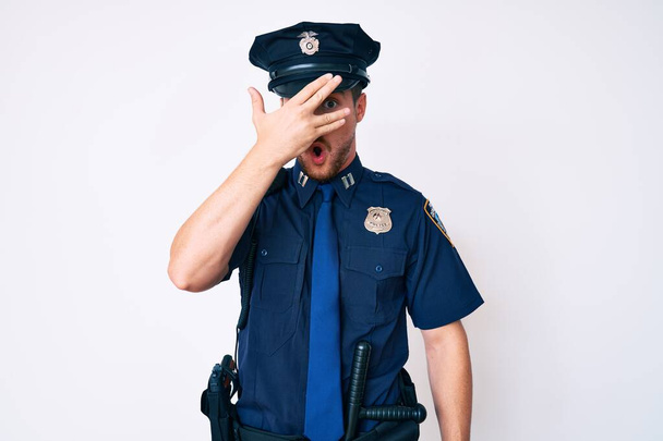 Giovane uomo caucasico indossa uniforme della polizia sbirciare in shock che copre il viso e gli occhi con la mano, guardando attraverso le dita con espressione imbarazzata.  - Foto, immagini