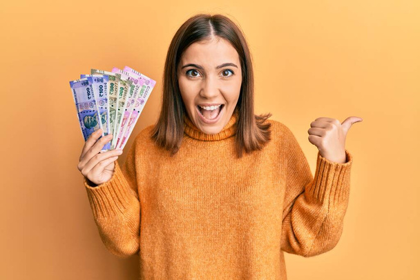 Young beautiful woman holding indian rupee banknotes pointing thumb up to the side smiling happy with open mouth  - Foto, afbeelding