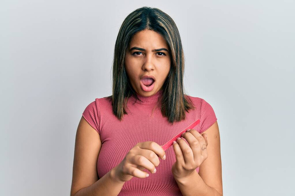 Jonge latijnse vrouw met behulp van bestand spijker in shock gezicht, op zoek sceptisch en sarcastisch, verrast met open mond  - Foto, afbeelding