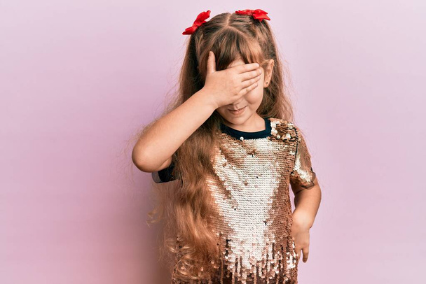 Malá kavkazská holčička, oblečená do slavnostních šatů, se usmívá a směje se s rukou na tváři, zakrývající oči pro překvapení. slepý koncept.  - Fotografie, Obrázek