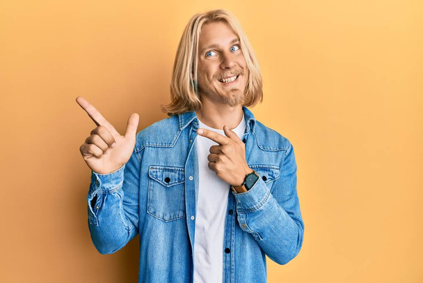 Homme caucasien aux cheveux longs blonds portant une veste décontractée en denim souriant et regardant la caméra pointant avec deux mains et doigts sur le côté.  - Photo, image