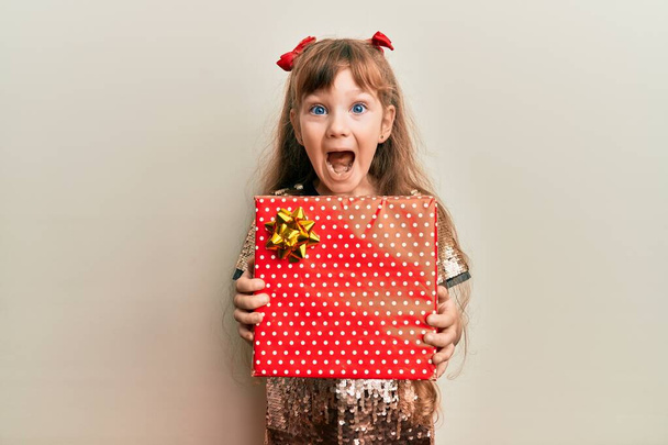 Μικρή καυκάσιος κορίτσι κρατώντας κόκκινο δώρο γιορτάζει τρελό και έκπληκτος για την επιτυχία με ανοιχτά μάτια ουρλιάζοντας ενθουσιασμένος.  - Φωτογραφία, εικόνα