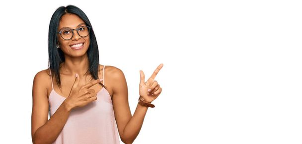 Νεαρή Αφροαμερικανή γυναίκα που φοράει καθημερινά ρούχα και γυαλιά χαμογελώντας και κοιτάζοντας την κάμερα με τα δύο χέρια και τα δάχτυλα στο πλάι.  - Φωτογραφία, εικόνα