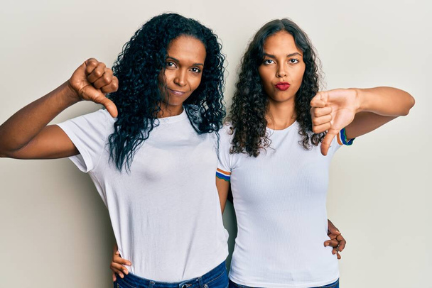 アフリカ系アメリカ人の母親と娘が怒った顔でカジュアルな白いTシャツを着て、否定的なサインは親指を下げて嫌いを示す、拒否の概念  - 写真・画像