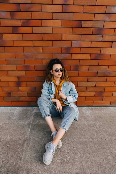 Mujer joven que usa ropa de mezclilla, sentada junto a una pared de ladrillo, al aire libre. - Foto, Imagen