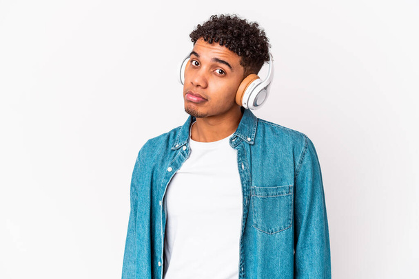 Genç Afro-Amerikan kıvırcık adamı, izole edilmiş kulaklıklı müzik dinliyor omuz silkiyor ve gözleri açık.. - Fotoğraf, Görsel