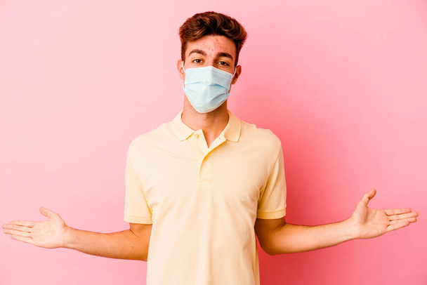 Joven hombre caucásico usando una protección para coronavirus aislado sobre fondo rosa que muestra una expresión bienvenida. - Foto, Imagen