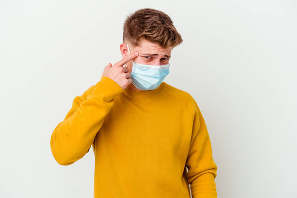 Hombre joven con una máscara para coronavirus aislado sobre fondo blanco que muestra un gesto de decepción con el dedo índice. - Foto, Imagen
