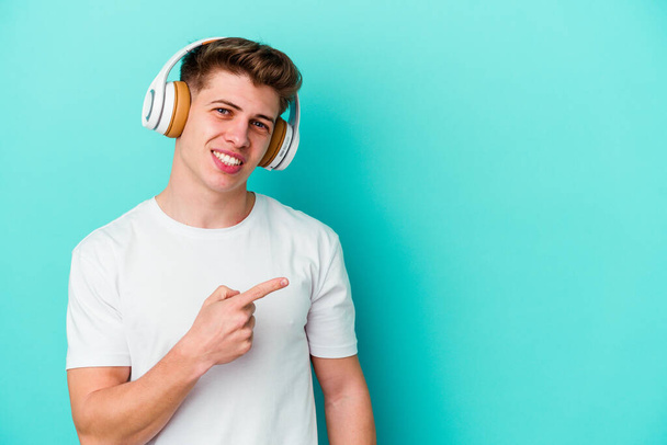 Jonge blanke man die naar muziek luistert met een koptelefoon geïsoleerd op een blauwe achtergrond die glimlacht en opzij wijst, en iets op een lege ruimte laat zien.. - Foto, afbeelding
