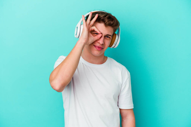 Jeune homme caucasien écoutant de la musique avec des écouteurs isolés sur fond bleu excité garder ok geste sur oeil. - Photo, image