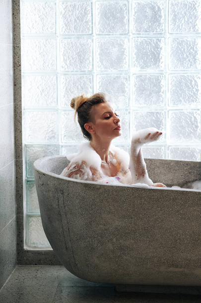 Красивая чувственная молодая женщина сидит в ванной и играет с пеной - Фото, изображение