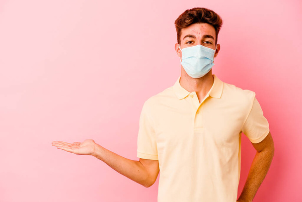 Junger kaukasischer Mann trägt einen Coronavirus-Schutz isoliert auf rosa Hintergrund, der einen Kopierraum auf einer Handfläche zeigt und eine andere Hand auf der Taille hält. - Foto, Bild