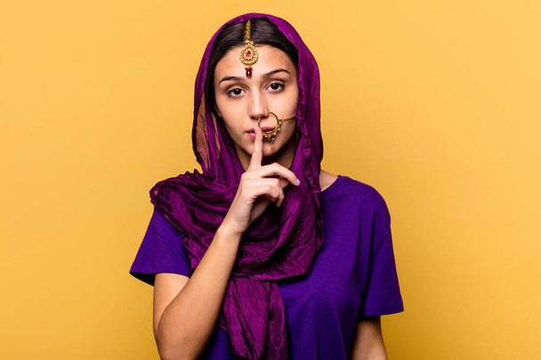 Nuori intialainen nainen yllään perinteinen sari vaatteita eristetty keltaisella taustalla pitää salaisuuden tai pyytää hiljaisuutta. - Valokuva, kuva