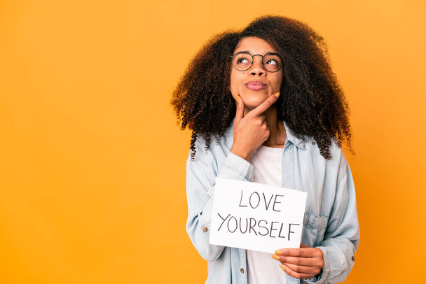 Jovem afro-americana encaracolado mulher segurando um amor você mesmo cartaz olhando para os lados com expressão duvidosa e cética. - Foto, Imagem
