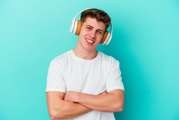 Jonge blanke man luisteren naar muziek met koptelefoon geïsoleerd op blauwe achtergrond lachen en plezier hebben. - Foto, afbeelding