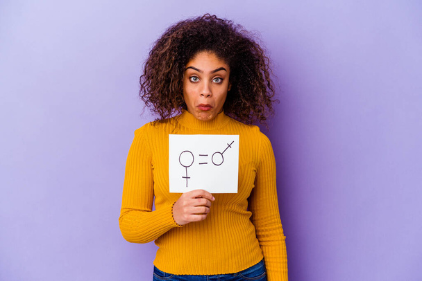 Young African American woman holding a gender equality placard isolated on purple background shrugs shoulders and open eyes confused. - Photo, Image