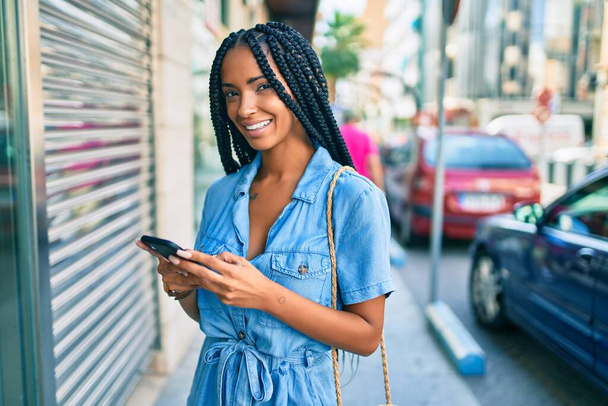 Mladý africký americký žena usměvavý šťastný pomocí smartphone ve městě. - Fotografie, Obrázek
