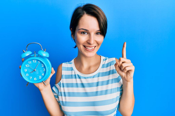 Young brunette woman with short hair holding alarm clock smiling with an idea or question pointing finger with happy face, number one  - 写真・画像
