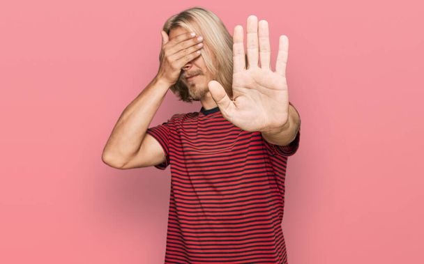 Kaukasischer Mann mit blonden langen Haaren, lässig gestreiftes T-Shirt, das die Augen mit Händen bedeckt und mit traurigem und ängstlichem Gesichtsausdruck Stopp-Gesten macht. Peinliches und negatives Konzept.  - Foto, Bild