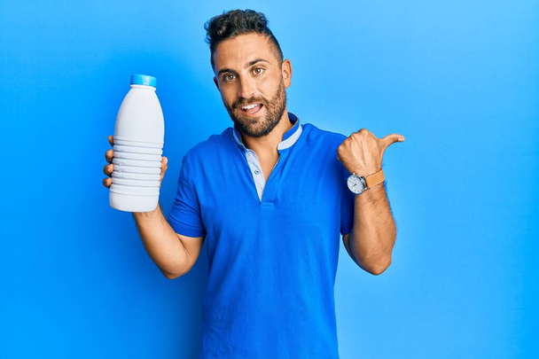 Handsome man with beard holding liter bottle of milk pointing thumb up to the side smiling happy with open mouth  - Foto, immagini
