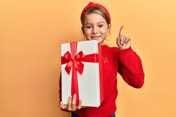 Little beautiful girl holding gift smiling with an idea or question pointing finger with happy face, number one  - Фото, изображение