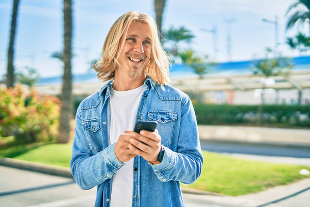 Молодий блондин скандинавський чоловік посміхається щасливим, використовуючи смартфон у місті
. - Фото, зображення