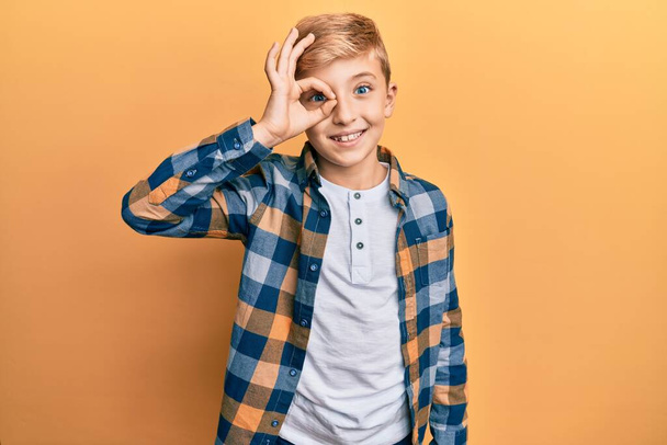 Pequeno garoto caucasiano vestindo roupas casuais sorrindo feliz fazendo ok sinal com a mão no olho olhando através dos dedos  - Foto, Imagem
