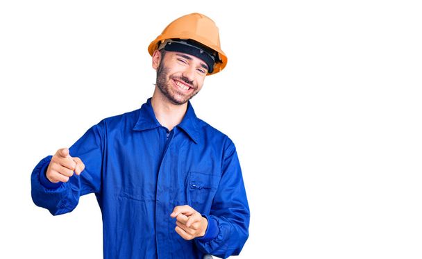 Een jonge Spaanse man in arbeidersuniform die met zijn vingers naar de camera wijst met een vrolijk en grappig gezicht. goede energie en vibes.  - Foto, afbeelding
