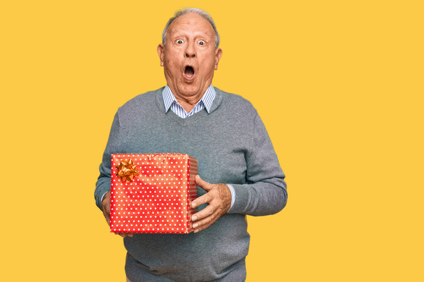 Старший кавказский мужчина держит в руках подарок испуганный и удивленный с открытым ртом для удивления, неверие лицо  - Фото, изображение