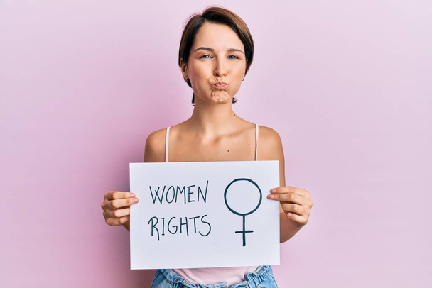 Young brunette woman with short hair holding woman rights banner puffing cheeks with funny face. mouth inflated with air, catching air.  - Photo, Image