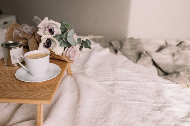 Mañana romántica. Mesa de centro de madera con flores en la cama con cuadros, taza de café, flores y velas. Rosas lila con eucalipto y anémonas. Tonos grises interiores. Foto de alta calidad - Foto, imagen
