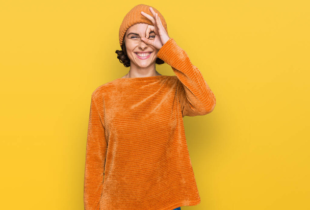 Młoda Latynoska kobieta w luźnych ubraniach i wełnianej czapce robi ok gest z uśmiechem dłoni, oko patrząc przez palce ze szczęśliwą twarzą.  - Zdjęcie, obraz