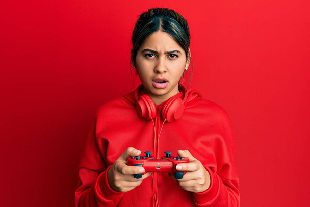 Video oyunu oynayan genç Latin kadın şok suratlı, şüpheci ve alaycı görünüyor, ağzı açık kalınca şaşırıyor.  - Fotoğraf, Görsel