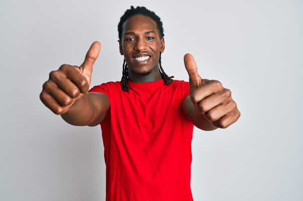 Αφροαμερικάνος με κοτσίδες που φοράει καθημερινά ρούχα εγκρίνοντας να κάνει θετική χειρονομία με το χέρι, με τους αντίχειρες σηκωμένους χαμογελαστούς και χαρούμενος για την επιτυχία. νικήτρια χειρονομία.  - Φωτογραφία, εικόνα