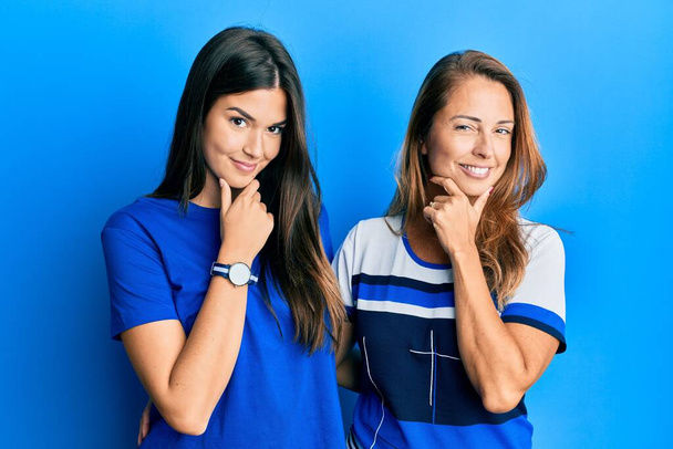 青い背景の上にカジュアルな服を着た母親と娘のヒスパニック系の家族は、カメラが交差した腕と手であごに提起笑みを浮かべて自信を持って見えます。ポジティブな.  - 写真・画像