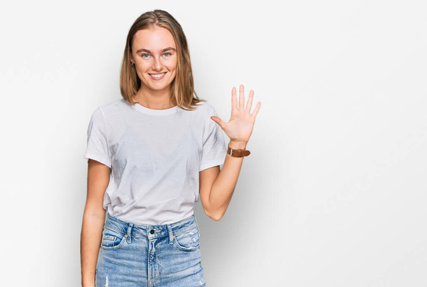 Belle jeune femme blonde portant un t-shirt blanc décontracté montrant et pointant vers le haut avec les doigts numéro cinq tout en souriant confiant et heureux.  - Photo, image