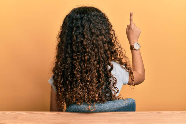 Adolescente hispana vestida con ropa casual sentada en la mesa posando hacia atrás apuntando hacia adelante con la mano del dedo  - Foto, Imagen