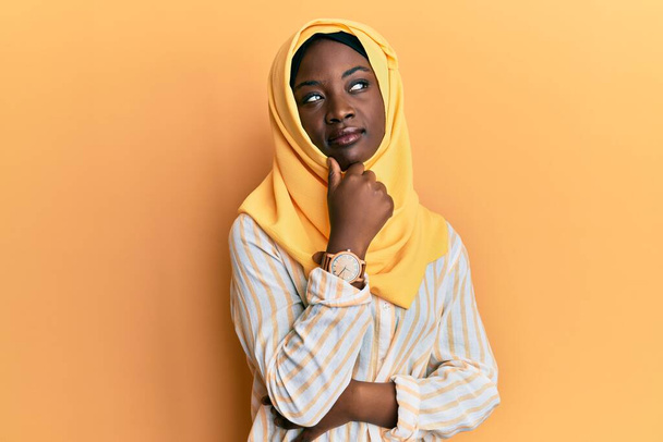 Όμορφη Αφρικανή νεαρή γυναίκα που φοράει παραδοσιακό ισλαμικό μαντήλι χιτζάμπ με το χέρι στο πηγούνι σκεπτόμενη την ερώτηση, την σκεπτική έκφραση. Χαμογελώντας με ευγενικό πρόσωπο. έννοια της αμφιβολίας.  - Φωτογραφία, εικόνα