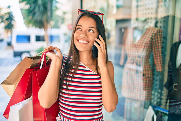 Fiatal latin vásárló lány gazdaság bevásárló táskák és beszél az okostelefon a városban. - Fotó, kép